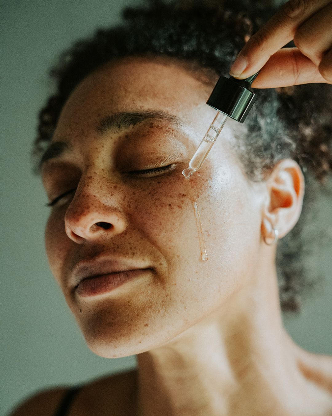 Woman holding dropper and dropping Finger Lime Face Lift Hydrating & Brightening Serum onto cheek.