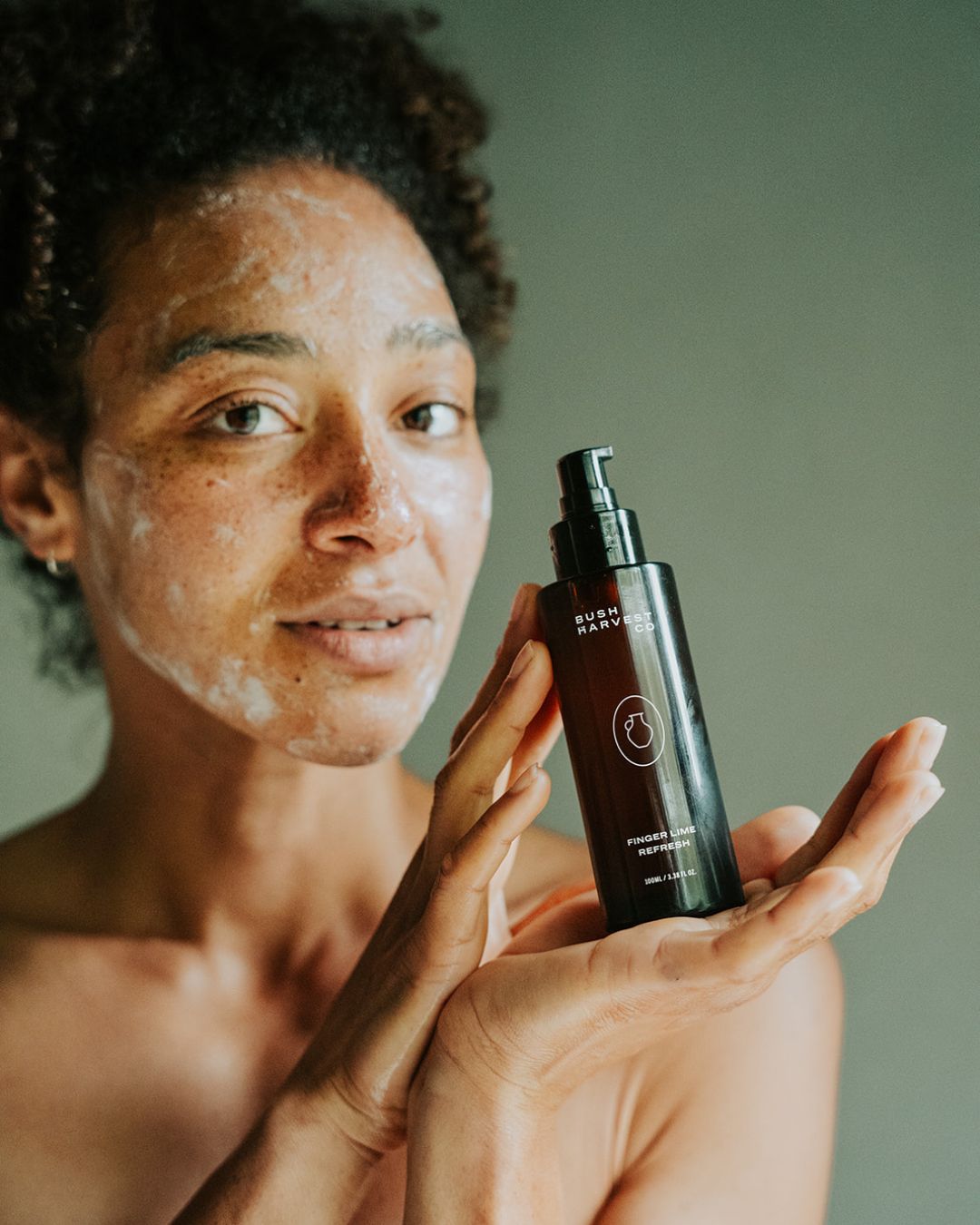 Woman with Finger Lime Refresh Purifying Facial Cleanser on face and holding product bottle.