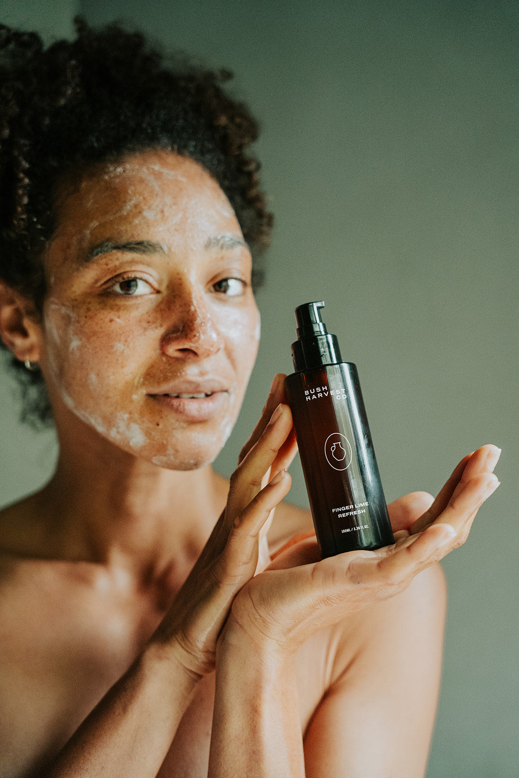 Woman with cleanser on her face holding Finger Lime Refresh Purifying Facial Cleanser bottle.