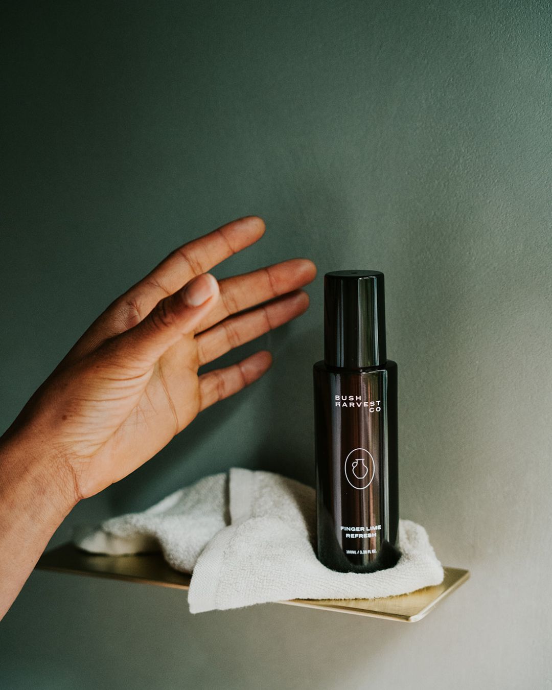 Woman's hand reaching for Finger Lime Refresh Purifying Facial Cleanser bottle, which is sitting with a reusable, bamboo cotton blend Facial Cleansing Cloth on a brushed gold shower shelf.